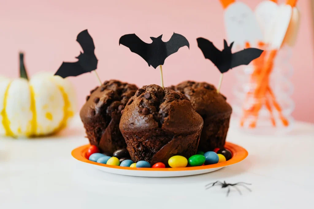 Czekoladowe muffiny z kawałkami czekolady i topperami w kształcie nietoperzy, na talerzyku z kolorowymi drażetkami, dekoracja na Halloween