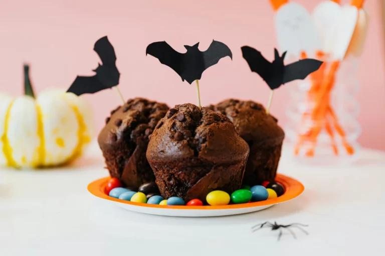 Czekoladowe muffiny z kawałkami czekolady i topperami w kształcie nietoperzy, na talerzyku z kolorowymi drażetkami, dekoracja na Halloween